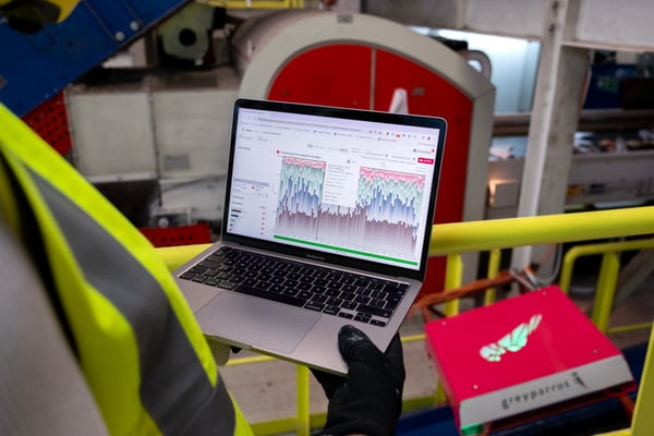 A recycling professional at a plastics recovery facility holding a laptop displaying the Greyparrot Analyzer portal, with a Greyparrot Analyzer unit in the background.